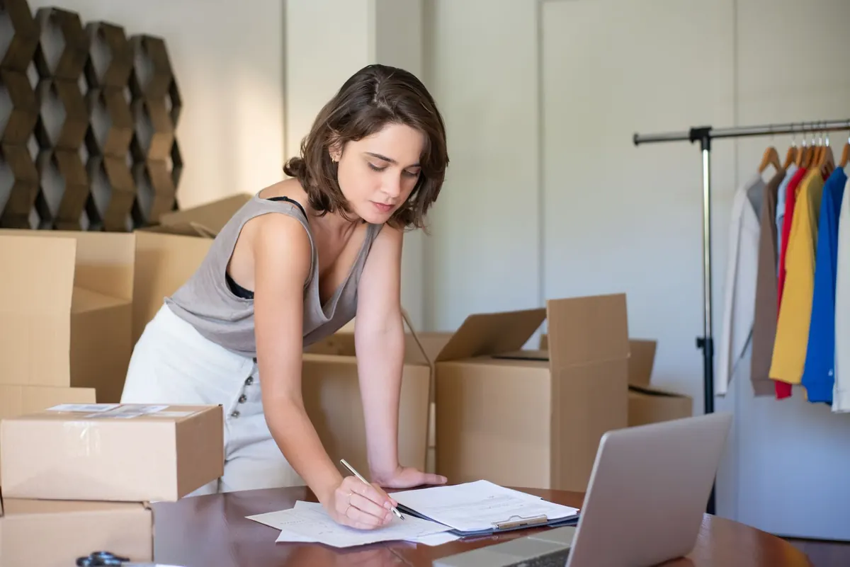 A woman leaning over a desk writing on a clipboard beside a laptop, surrounded by cardboard boxes and a rail of clothes.