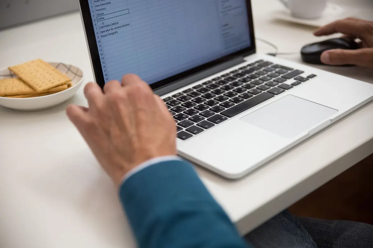 A laptop on a white desk showing a spreadsheet with a column of names selected, a mouse and a bowl of crackers beside it.