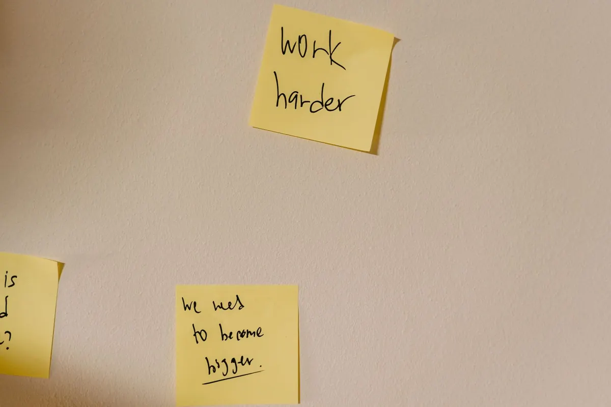 Yellow sticky notes on a plain wall, one reading work harder and another reading we need to become bigger.