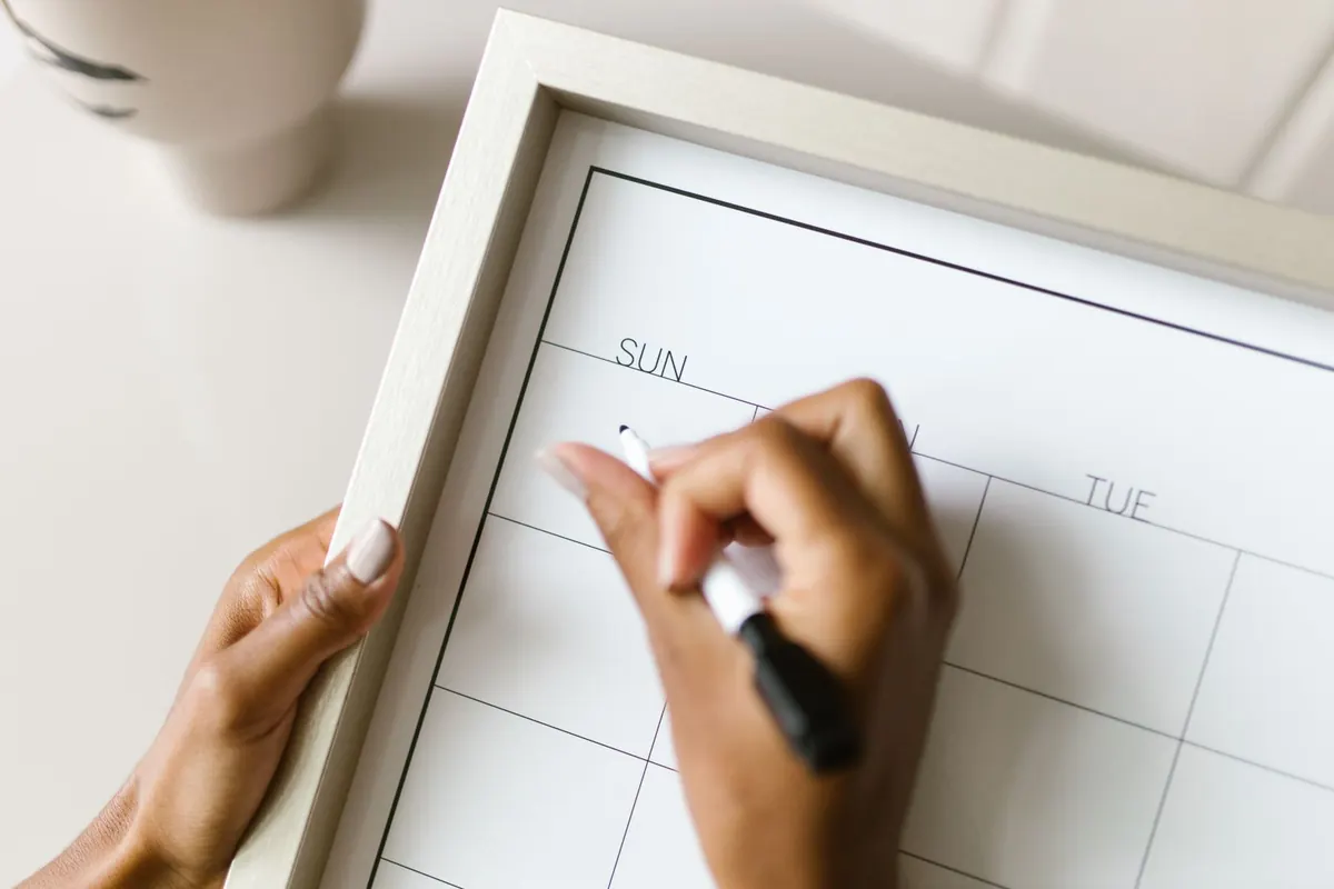 Hands holding a framed whiteboard planner marked with the days of the week, writing in one of the empty boxes.