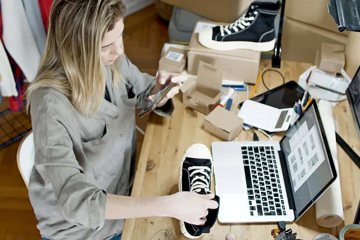 A woman photographing a trainer with her phone at a desk covered in shipping boxes, a laptop open beside her.