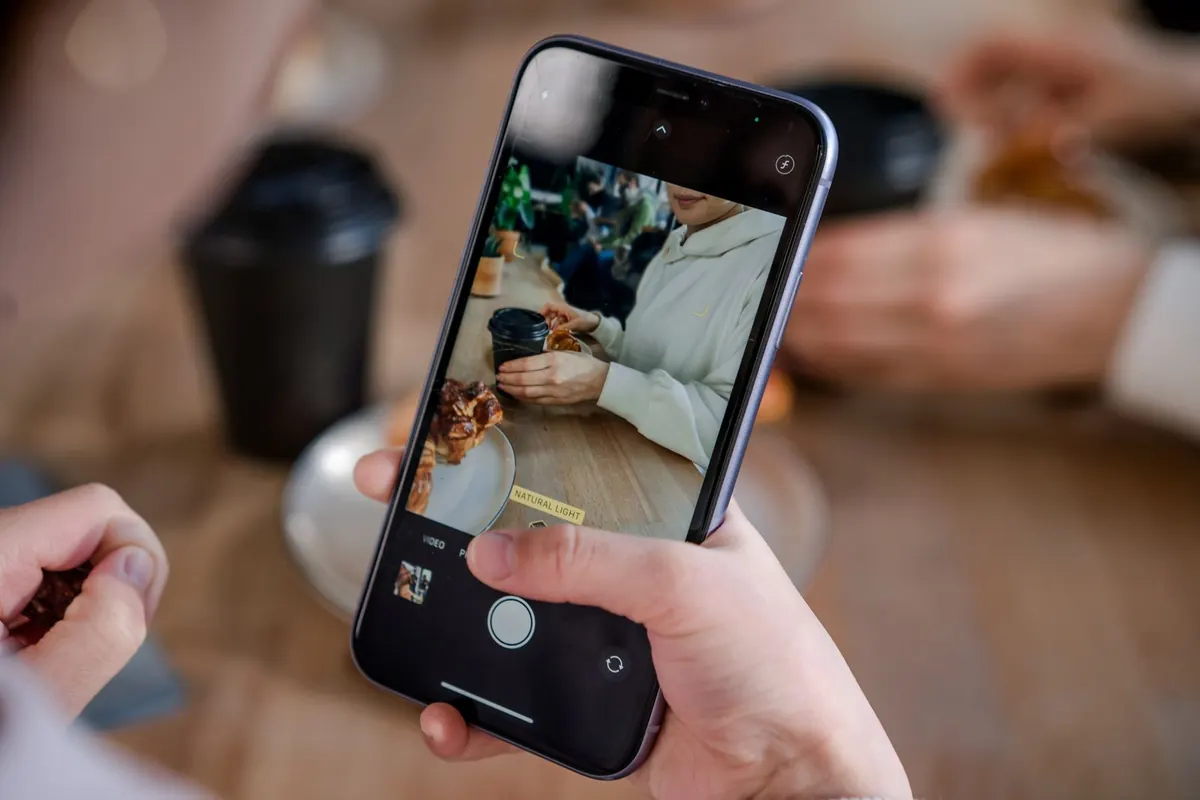 A hand holding a phone in camera mode, filming someone across a cafe table with a coffee and a pastry.