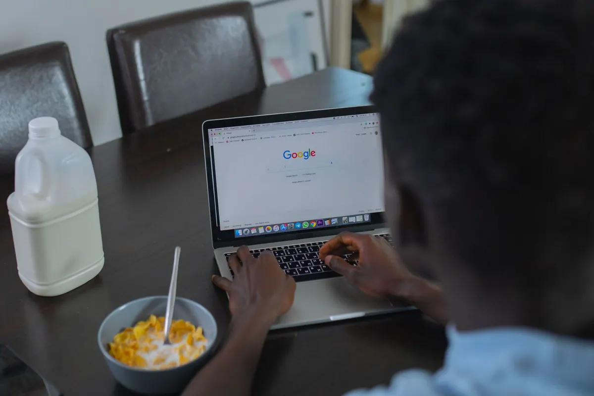 Someone at a kitchen table with a bowl of cereal, a laptop open on a search page.