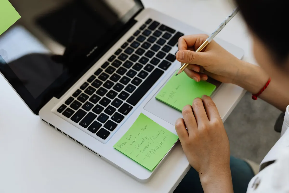 Someone writing on green sticky notes stuck to a laptop.