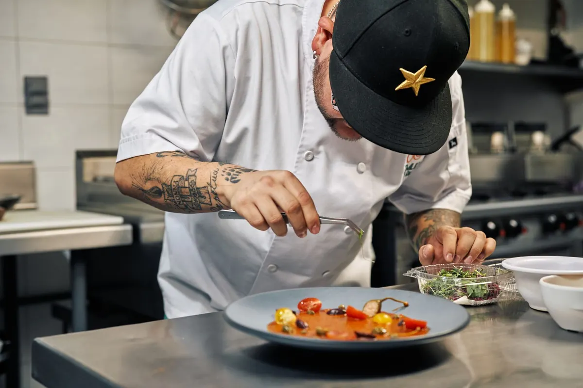 A chef in whites using tweezers to garnish a plate in a stainless steel kitchen.