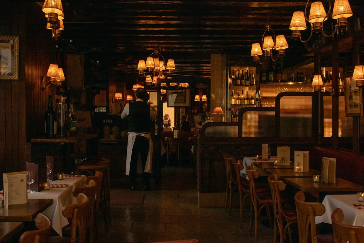 A dim restaurant interior lit by small warm lamps, with laid tables and a waiter at the bar.