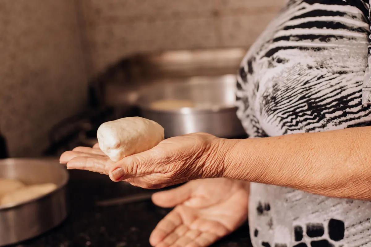 An older cook's hands holding a ball of dough beside steel pans in a kitchen.