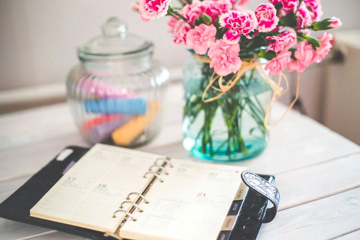 An open paper planner on a desk beside a vase of pink flowers.